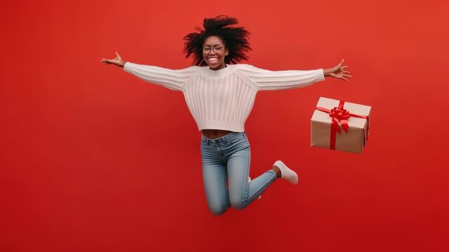 Excited woman jumping with a gift box in a red background. Overjoyed young girl with curly hair is excited with the present. Happiness and holidays concept. 