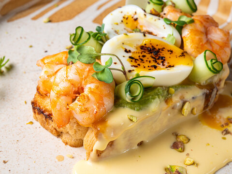 A gourmet open-faced toast topped with grilled shrimp, soft-boiled egg, avocado spread, cucumber ribbons