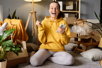 A woman in a yellow hoodie sits cross-legged on the floor amidst moving boxes, holding a smartphone...