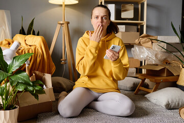A woman sits on the floor among moving boxes, looking shocked at her phone after moving into her new home.