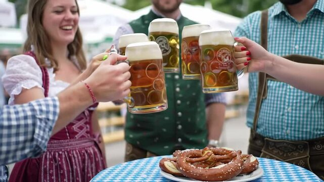 A group of friends, including a woman and men, in traditional dress clinking full beer mugs, celebrating Oktoberfest with pretzels on a checkered tablecloth, cheers footage.