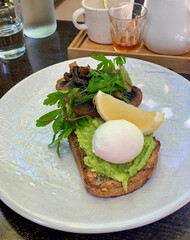A beautifully presented avocado toast topped with a perfectly poached egg, sautéed mushrooms, fresh herbs, and a lemon wedge, served on a light-colored plate.