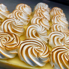 A close-up view of a vibrant lemon meringue pie or tart, featuring beautifully piped and toasted meringue peaks over a bright yellow lemon custard filling.
