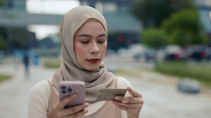 Portrait of young Malaysian woman wearing headscarf pays online uses credit card and smartphone on street. Muslin female inserts banking data to transfer money via app outdoors - Powered by Adobe