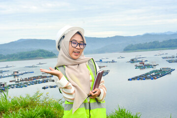 A female fish farm manager wears a safety vest and carries a book to check the work.