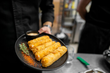 A chef presents black plate with four crispy breaded sticks, garnished with thyme and red pepper flakes, accompanied by small bowl of dipping sauce. background features a professional kitchen setting