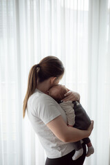 Young mother holding newborn baby in soft natural light standing by a window. Concept of early...