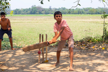 A village boy plays a shot during a game of cricket on a dusty field, while another boy watches intently in the background with open farmland stretching behind them