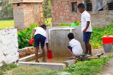 Three schoolboys in uniform gather around a small outdoor water station, filling red buckets as...