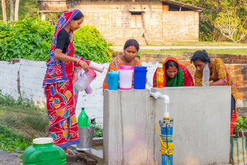 A group of women in colorful sarees gather around a community tap, filling containers with water...