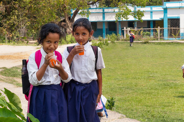 Two schoolgirls in uniform enjoy orange drinks with straws, smiling with backpacks on their shoulders