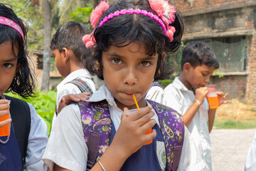 A young schoolgirl with a pink headband looking at the camera while sipping an orange drink