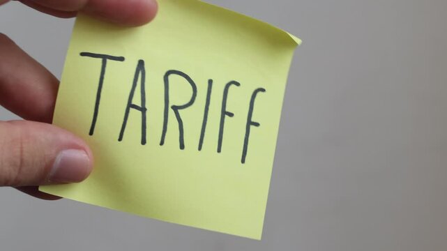 A hand holds a bright yellow sticky note with the word "TARIFF" written in bold black letters, against a plain background. Ideal for news, economics, business, or political content