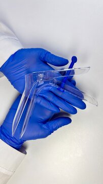 Medical professional's hands in blue gloves holding a transparent plastic speculum against a neutral background. The image conveys sterile handling examination equipment