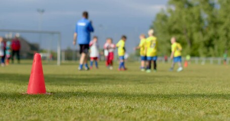 Children in bright yellow and red soccer uniforms practice on a wellkept grass field, with colorful cones around, as a coach supervises. Goal posts indicate its soccer practice - Powered by Adobe