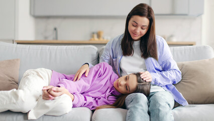 Loving mother comforting and talking to her teen daughter, tired upset girl lying on mom knees, sitting on sofa at home