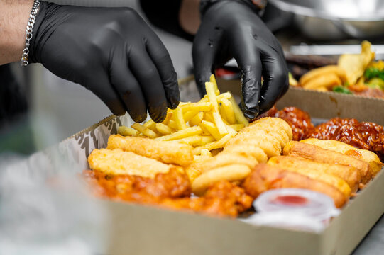 A person wearing black gloves arranges crispy fried foods like French fries and chicken tenders in a takeout container. The image showcases fast food preparation and presentation