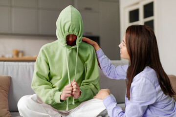 Upset teen girl wearing hood ignoring talk with mother, sitting on couch. Teenager introvert hiding...