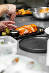A chef's hand garnishes a plated dish with grilled chicken, pickles, and sauce on a black platter in a professional kitchen. The scene highlights culinary precision and food presentation