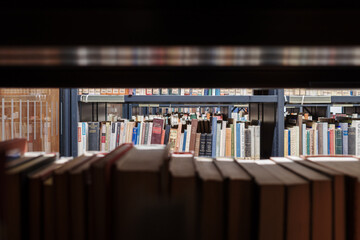 A view from between two dark shelves in a library or archive. The foreground shows a row of old,...