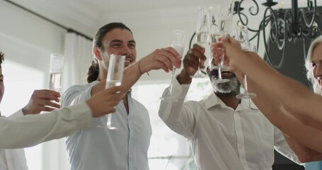 Diverse friends toasting with flutes under chandelier, clinking, sipping and laughing at home - Powered by Adobe