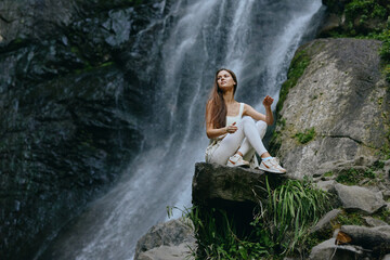 young woman in white clothes sitting on rock near waterfall surrounded by lush greenery in natural...
