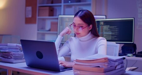 Close up of Stressed Asian woman works overtime in dark modern office filled with paper stacks, monitors, and code screens. Exhausted female employee under pressure to meet tight deadline alone.