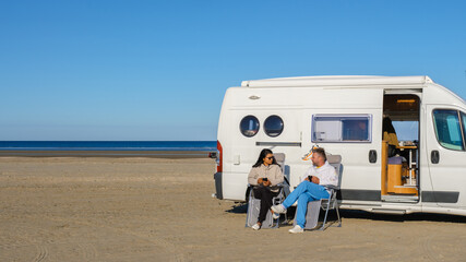 Two people relax by a white camper van parked on the sandy beach of Lakolk Strand, Romo Denmark, enjoying the warm sun and beautiful coastline. Ideal for travel and outdoor lifestyle themes.