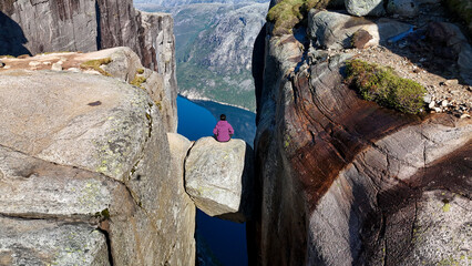 An adventurous individual sits on a massive boulder suspended between two steep cliffs, surrounded...