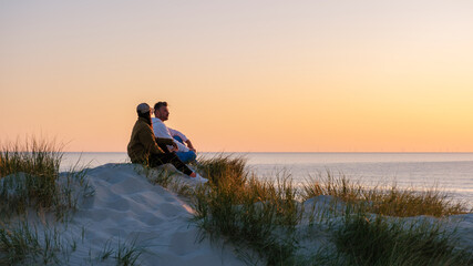 Friends relaxes on soft sand dunes while watching a breathtaking sunset over the tranquil waters of...