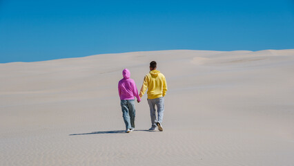 young couple dressed in colorful hoodies strolls hand in hand across sandy dunes against a vibrant...