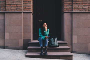 Sidewalk scene with young woman seated near doorway, holding planner and smartphone, looking into...