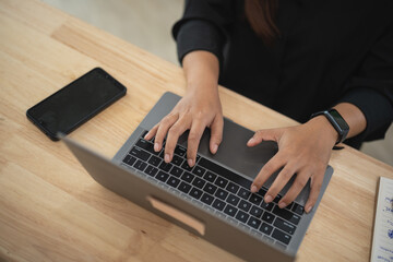 Close-Up of Hands Typing on Laptop Keyboard with Smartphone Nearby on Wooden Table in Bright Office Environment, Showcasing Modern Work Culture