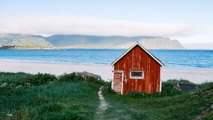 A charming red cabin stands near the sandy beach in Lofoten Norway, framed by lush greenery and distant mountains. The clear blue waters stretch to the horizon, offering a peaceful coastal retreat.