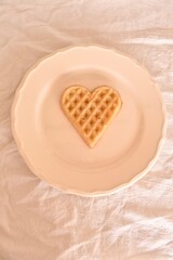 Single heart-shaped Belgian waffle on a white plate. Simple and soft breakfast or dessert with clean presentation.