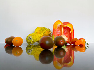 A vibrant display of fresh vegetables and fruits including a yellow squash, red bell pepper, and various tomatoes rests on a glossy surface, showcasing their colors and reflections.