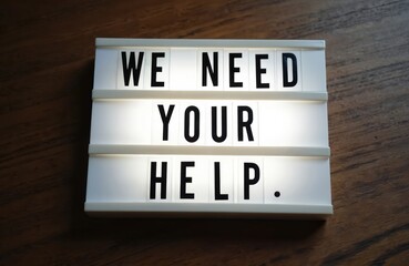 Light box on wooden background. Text message We Need Your Help. Symbol of support. Cooperation, charity, giving, communication. Concept of teamwork, assistance for business, donation, volunteering.