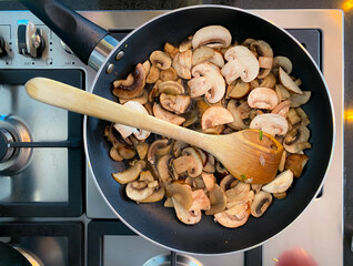 Fresh mushrooms are sliced and saut&eacute;ed in a skillet on a sleek stovetop. A wooden spoon is used to stir the mushrooms as they cook, highlighting the culinary process.