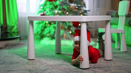 Caucasian toddler in Santa cap lies on the floor hiding behind the desk. Funny baby boy plays at home for Christmas. - Powered by Adobe
