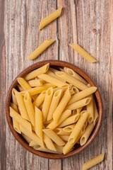 Top view of dry penne pasta in a wooden bowl with a few scattered pieces on aged wooden background. Warm food styling, natural texture, and rustic atmosphere.