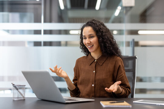 A businesswoman happily engaged in a video call, gesturing while sitting at her office desk. The laptop, notebook and tablet are on the table.