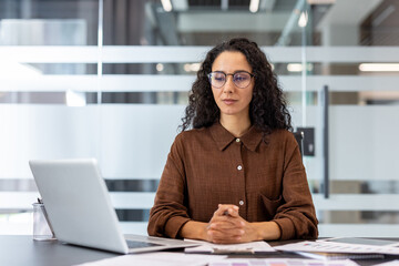 A woman in glasses sits at a desk, looking at a laptop and documents in a modern office setting.