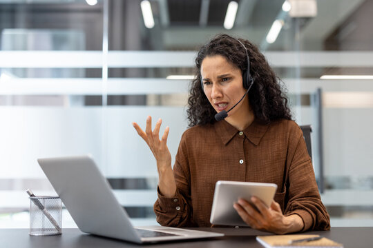 Anxious woman wearing a headset has an angry expression while working on a computer and a tablet. She is frustrated.