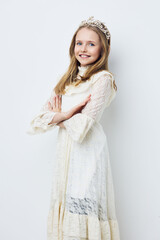 Happy young girl in a white lace dress and crown, posing confidently against a light background,...