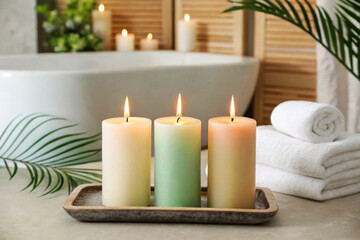 Three lit candles in soft colors on a tray, surrounded by towels and green leaves. A serene bathroom setting with a bathtub in the background.