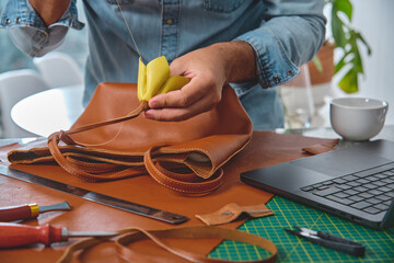 Leather craftsman is sewing a bag handle in his workshop, using traditional techniques and...