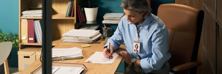 Middle aged Caucasian woman social worker sitting at office desk reviewing client documents and...