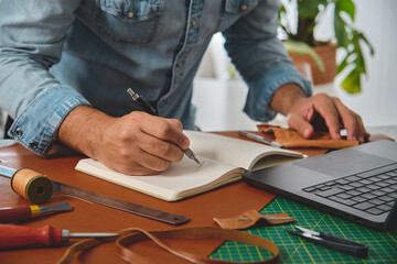 Shoemaker sketching a new model in a notebook, surrounded by leather crafting tools and a laptop