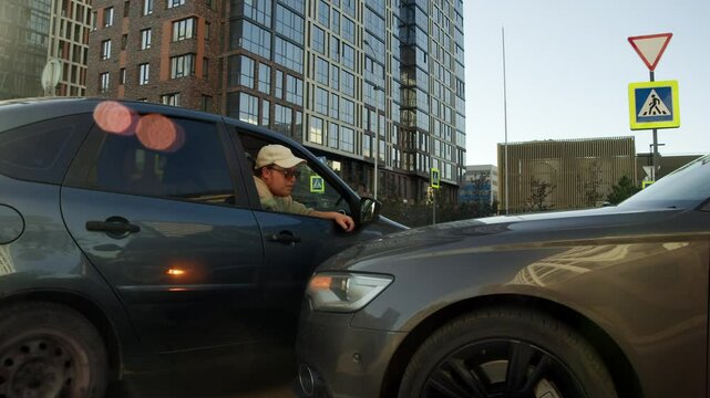 Bearded man, leaning out of window of his car, is arguing with careless driver who has just crashed into his car while driving in city.