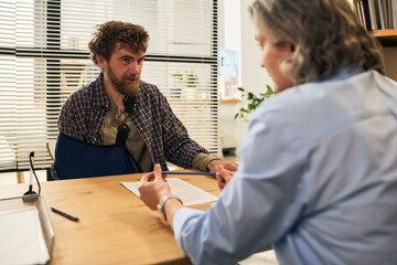 Middle aged Caucasian man with beard and arm sling sitting across desk from social worker discussing paperwork in bright office setting